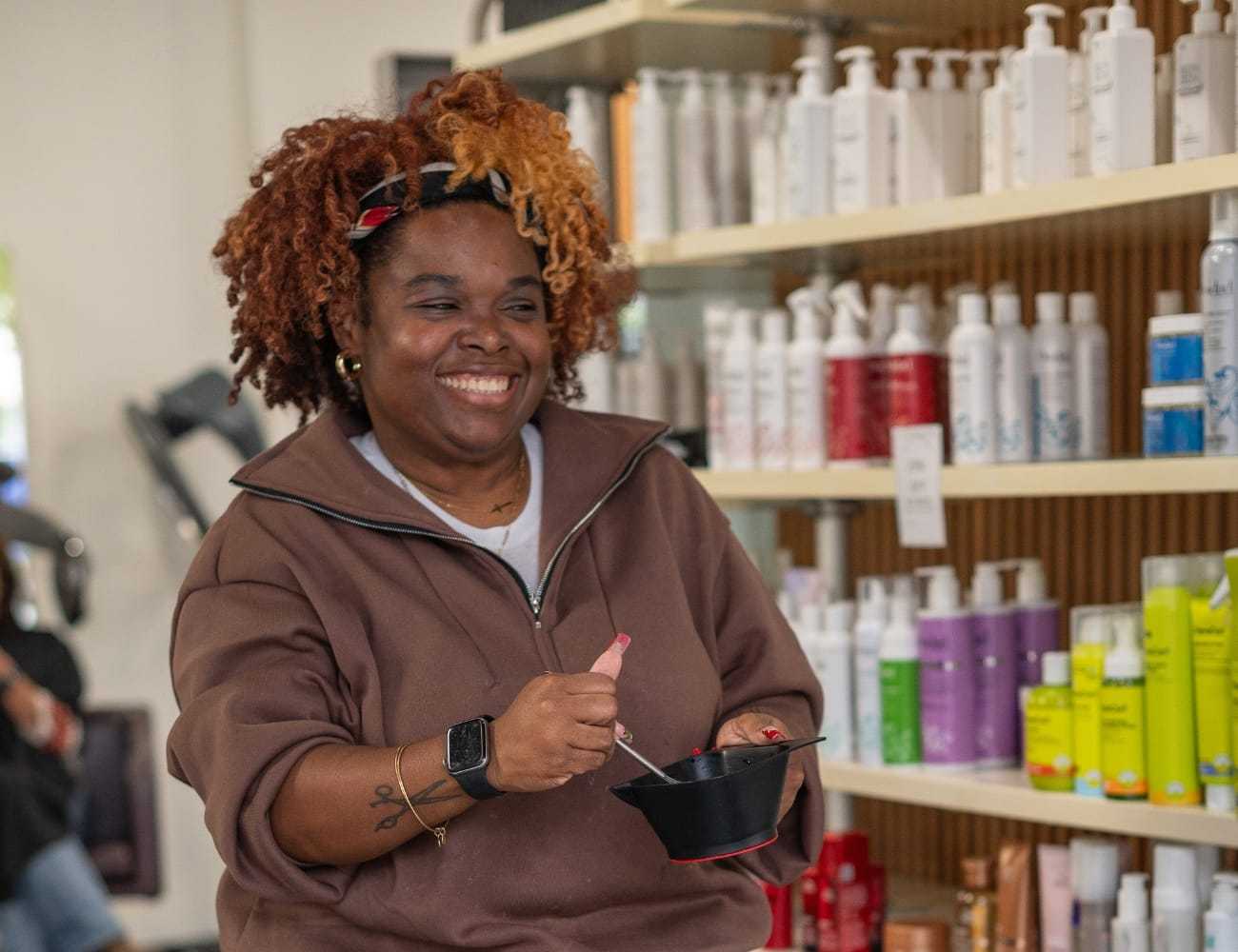 Smiling person preparing hair dye in salon, surrounded by colorful product bottles.