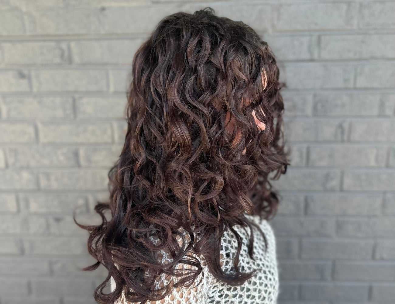 Curly-haired person facing a gray brick wall, wearing a light knit sweater.