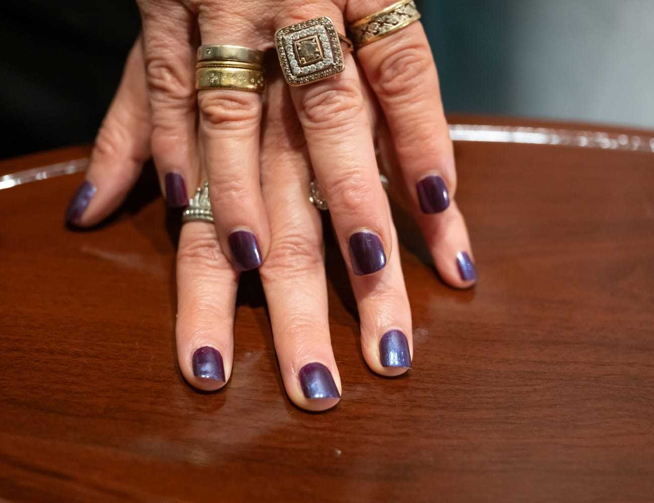 Purple manicured nails and silver rings on a wooden table surface.