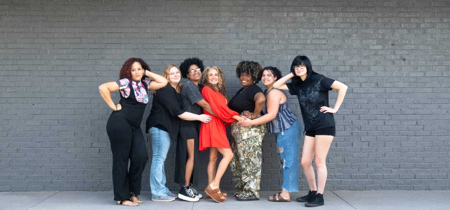 Seven women posing together in front of a gray brick wall, smiling and supporting each other.