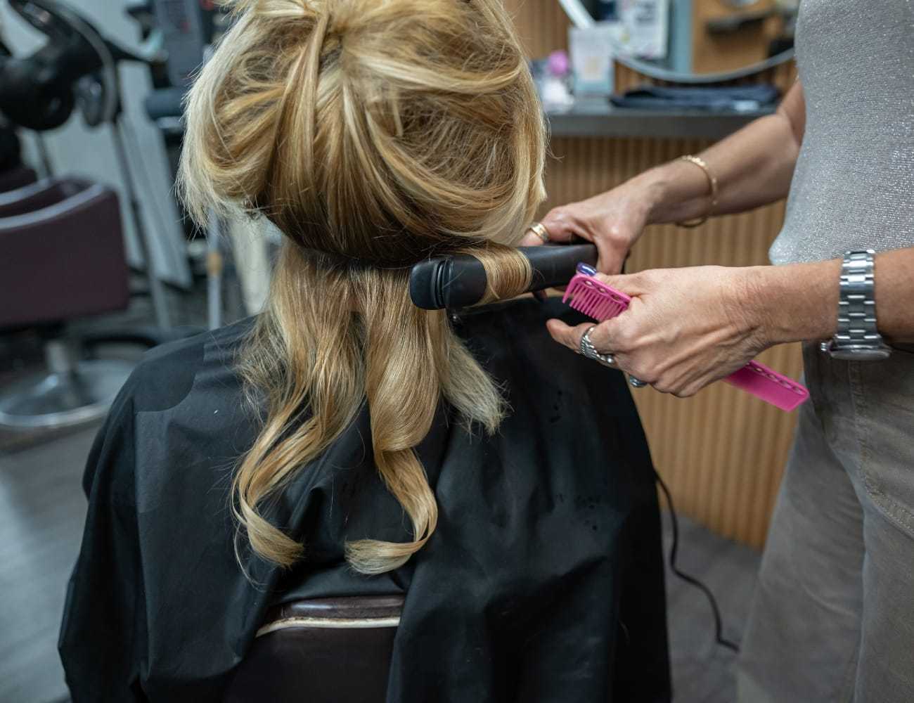 Stylist curling a woman's hair in a salon using a curling iron and pink comb.