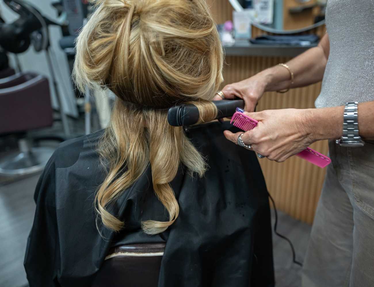 Stylist curling a woman's hair in a salon using a curling iron and pink comb.
