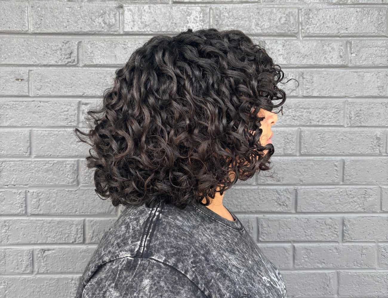 Curly-haired person in profile against a gray brick wall, wearing a dark textured shirt.